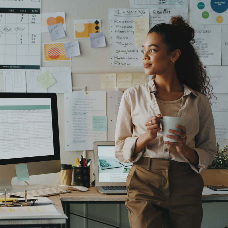 Confident woman holding coffee mug next to her computer with notes on the wall