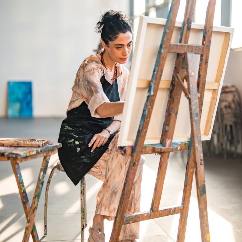 A young Middle Eastern female artist, wearing a paint-stained apron, paints on a canvas in a bright, naturally lit art studio, surrounded by easels and art supplies.