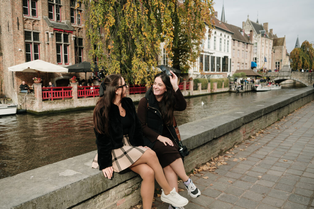 Two cheerful women in Bruges, Belgium sit next to a canal