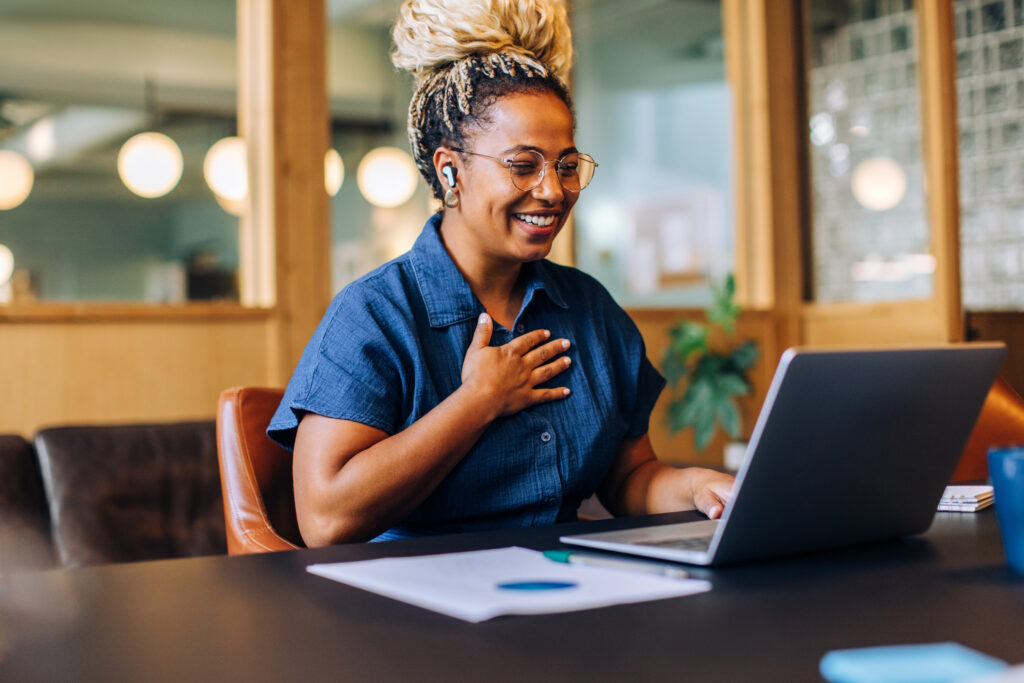 A cheerful young woman participates in an online meeting, showing lively engagement and joy. She sits at a desk in a bright room, using a laptop, with paperwork and a relaxed atmosphere.