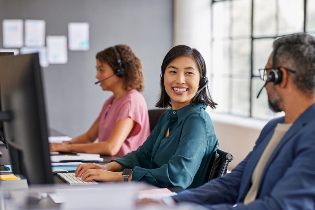 Happy customer service representative interacting with colleagues. Employee smiling while collaborating with support team. Young asian woman with headset talking to her coworker while working.